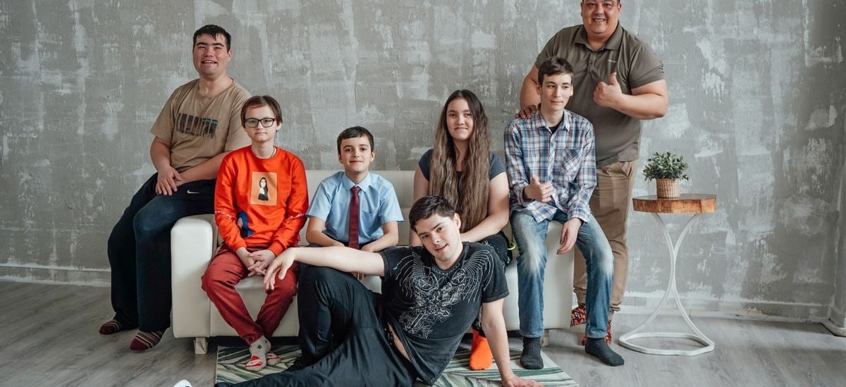 Group of seven people posing casually in a stylish indoor setting with a concrete wall background.